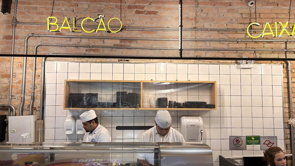 Balcão do Restaurante e Peixaria Pompéia: parede de azulejos brancos, tijolo aparente e letreiros em neon amarelo escritos Balcão e Caixa, com dois sushimen trabalhando